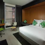 نمایی از اتاق Garden Classic با یک تخت دو نفره راحت و بالش‌های سبز رنگ، فضای کاری مدرن و نمای شهری از پنجره بزرگ.